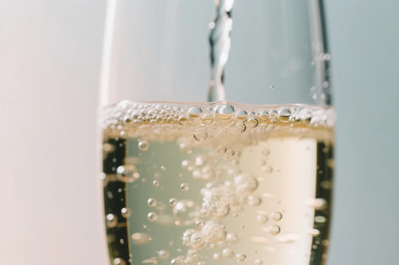 Een close-up toont mousserende wijn die in een glas wordt geschonken, met opstijgende belletjes in zacht licht en lichtgouden, witte en lichtblauwe tinten.