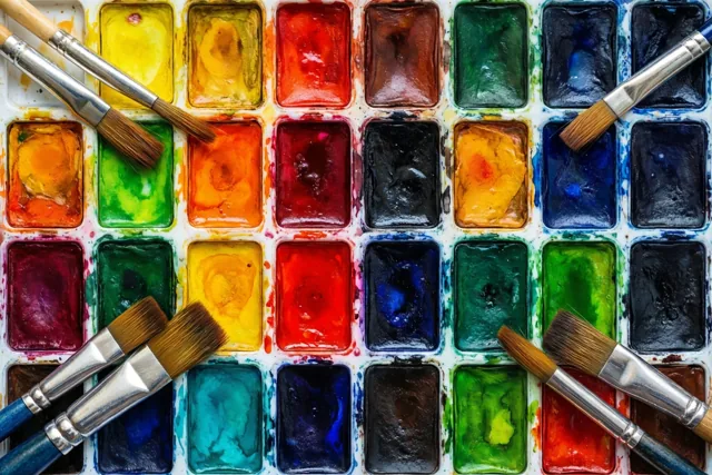 Op een aquarelpalet met rechthoekige vakjes vol gemengde rode, gele, groene, blauwe en zwarte verf liggen verschillende natte penselen, van bovenaf gezien.