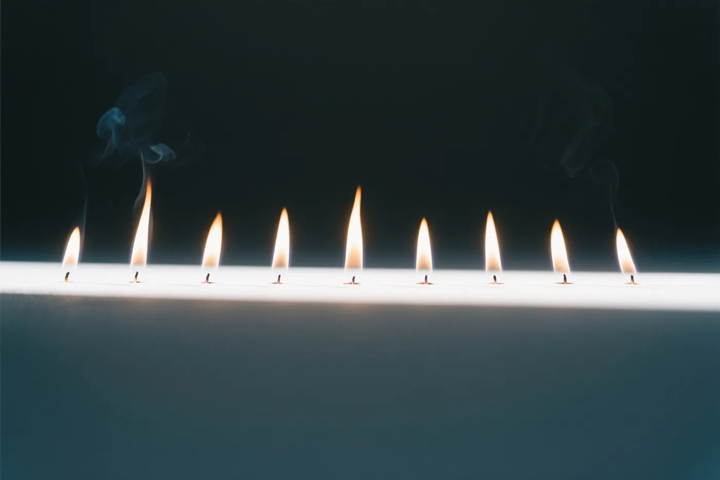 Negen kleine vlammen branden in een rechte lijn tegen een donkere achtergrond, met warm oranje licht en koele blauwgrijze tinten.