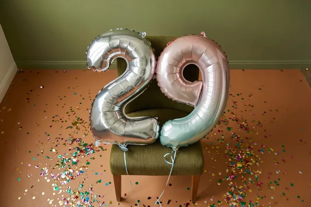 Zilveren en roségouden cijferballonnen in de vorm van 2 en 9 zijn vastgebonden aan een groene stoel, met confetti op een terracottavloer voor een gedempte groene muur.