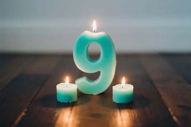 Een brandend theelicht in de vorm van een 9 staat tussen twee kleinere brandende theelichtjes op een donkere houten vloer, met een warme gloed en blauwgroene en bruine tinten.