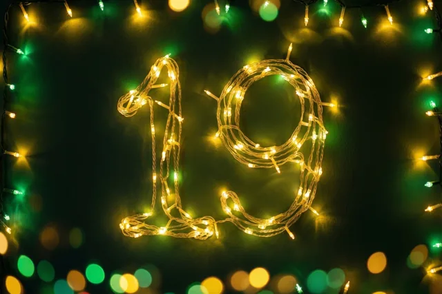 Warme lichtjes vormen 19 op een donkere muur, omringd door onscherpe feestlichtjes in gele en groene tinten.