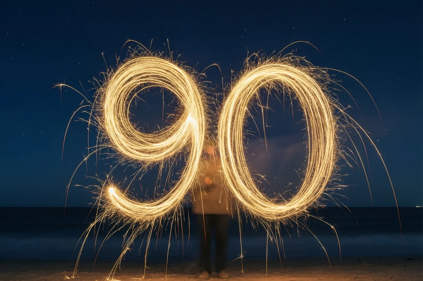 Een persoon schrijft 's nachts op een strand het getal 90 met gouden lichtsporen van sterretjes tegen een donkere hemel.