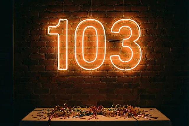 Een gloeiend neonbord met 103 hangt aan een donkere bakstenen muur boven een tafel met kleurrijke slingers in warm oranje en rood licht.