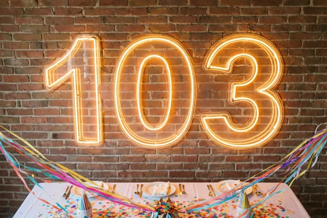 Een warm oplichtende neonreclame met 103 hangt aan een rode bakstenen muur boven een feesttafel met kleurrijke slingers en confetti.