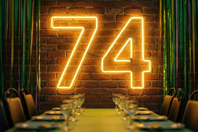 Een groot neongetal 74 straalt aan een bakstenen muur boven een gedekte feesttafel met groene en gouden slingers.