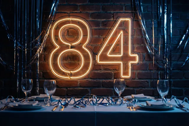 Een warm neonlicht in de vorm van 84 hangt aan een bakstenen muur boven een gedekte feesttafel met blauwe slingers.