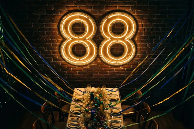 Een groot neongetal 88 hangt aan een bakstenen muur boven een feesttafel, met kleurrijke slingers en warm goudkleurig licht.