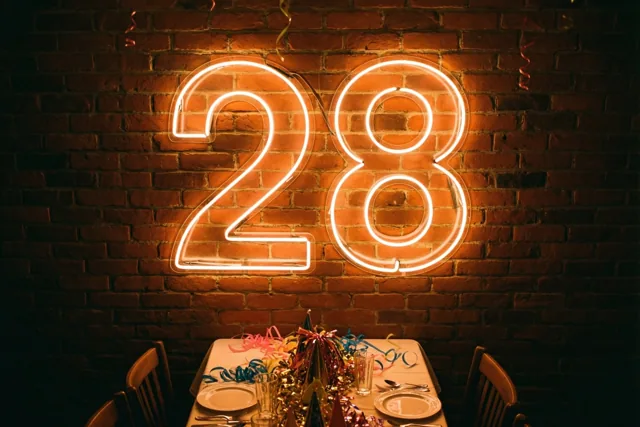 Een neonlicht in de vorm van 28 hangt aan een bakstenen muur boven een feesttafel met kleurrijke slingers en servies.
