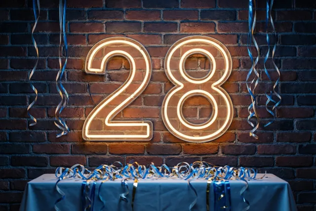 Een groot neonlicht in de vorm van 28 straalt tegen een donkere bakstenen muur boven een tafel met blauwe en zilveren slingers.