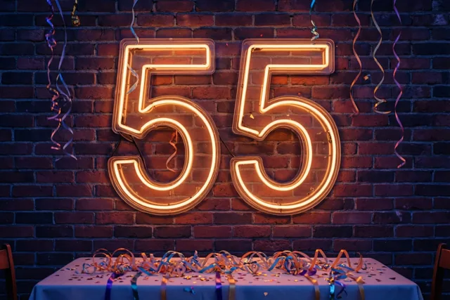 Een oranje neongetal 55 verlicht een donkere bakstenen muur boven een tafel met een lichtblauw tafelkleed, kleurrijke slingers en confetti.