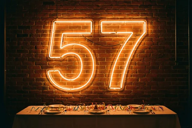 Een groot neonbord met 57 gloeit aan een bakstenen muur boven een feesttafel met borden, bestek en kleurrijke slingers.
