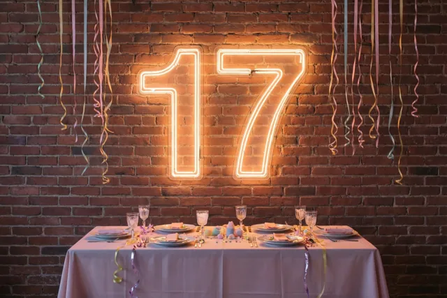 Een feesttafel met slingers staat voor een bakstenen muur met een groot neongetal 17 in warm roze, oranje en geel licht.