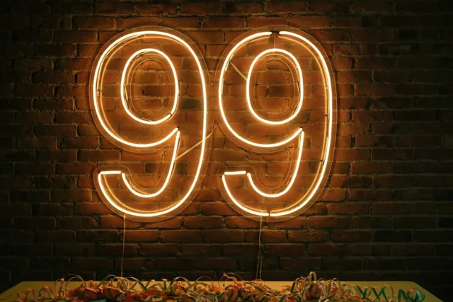 Een lichtgevend neonbord met 99 hangt aan een donkere bakstenen muur boven een tafel met een groen kleed en kleurrijke slingers.