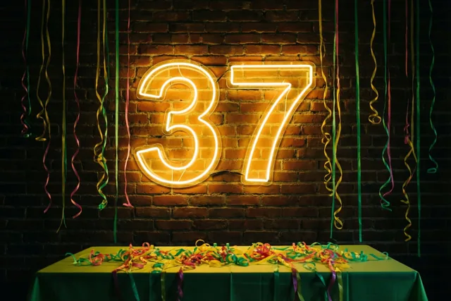 Een neonbord met 37 hangt aan een bakstenen muur boven een tafel met een groen kleed en slingers, met kleurrijke slingers erboven.