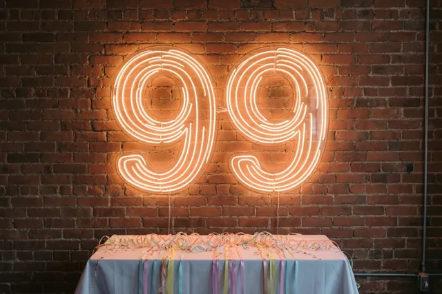 Een neonbord met 99 hangt aan een rode bakstenen muur boven een tafel met een lichtblauw kleed en kleurrijke slingers in warm licht.