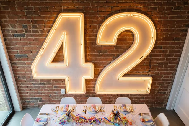 Een groot neongetal 42 verlicht een rode bakstenen muur boven een feestelijk gedekte tafel met kleurrijke slingers en confetti.