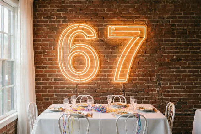 Een groot neongetal 67 verlicht een bakstenen muur boven een feesttafel met slingers.