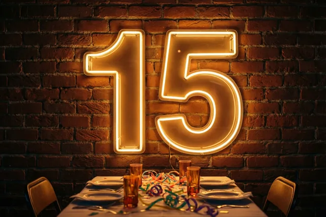 Een neongetal 15 hangt aan een bakstenen muur boven een feesttafel met borden, glazen en kleurrijke slingers in warm oranjegeel licht.