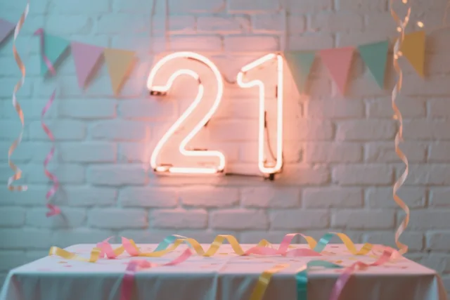 Een neonlicht met het getal 21 hangt boven een feesttafel aan een witte bakstenen muur, tussen kleurrijke slingers en driehoekige vlaggetjes.