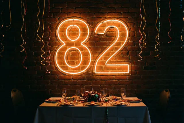 Een warm gloeiend neongetal 82 hangt aan een bakstenen muur boven een feesttafel met een wit tafellaken en hangende slingers.