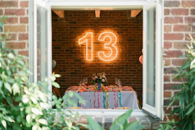 Door een open raam met groen op de voorgrond is een bakstenen muur met een lichtgevend neongetal 13 te zien boven een versierde tafel met een wit tafelkleed.