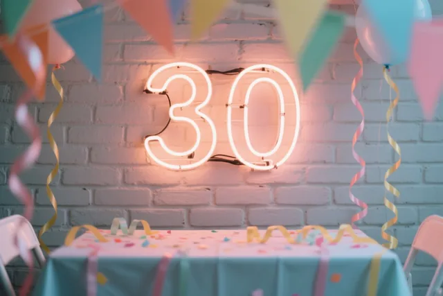 Een lichtgevend neongetal 30 hangt aan een witte bakstenen muur boven een feesttafel met confetti en serpentines, omringd door kleurrijke ballonnen en vlaggetjes.