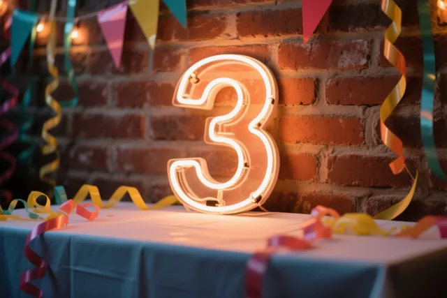 Een lichtgevende neon-3 hangt aan een bakstenen muur boven een tafel met een blauw tafelkleed en kleurrijke slingers, in warm licht.