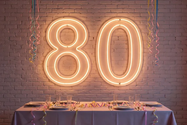 Een grote 80 in warm neonlicht hangt aan een bakstenen muur boven een gedekte feesttafel met gekleurde slingers.