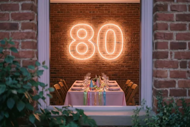 Door een raam is een lichtgevende 80 in neon aan een bakstenen muur te zien boven een feesttafel met slingers en glazen in roze en oranje licht.