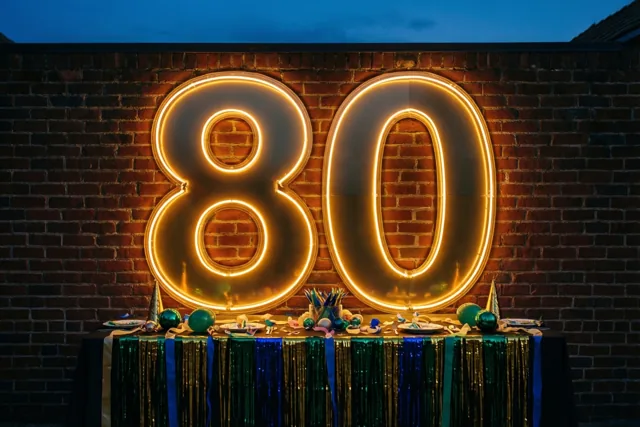Een grote 80 in warm neonlicht hangt aan een bakstenen muur boven een versierde feesttafel met groen-blauwe slingers, onder een blauwe hemel bij schemering.