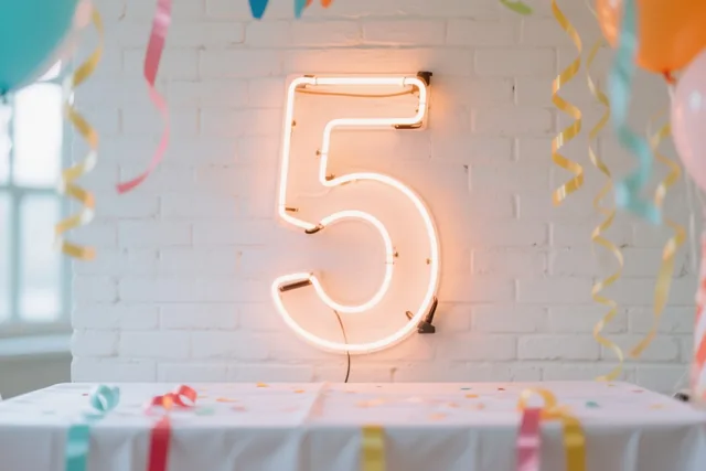 Een lichtgevend neoncijfer 5 hangt aan een witte bakstenen muur boven een feesttafel met kleurrijke slingers en ballonnen.