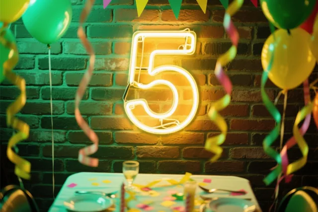 Een lichtgevend neoncijfer 5 hangt aan een bakstenen muur boven een versierde tafel met kleurrijke ballonnen en slingers in warme gele, groene en rode tinten.