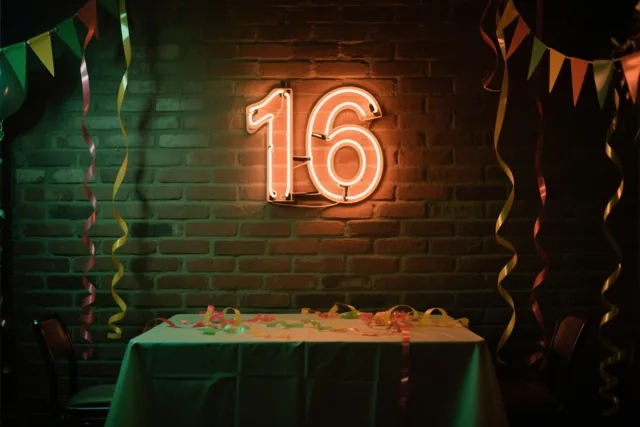 Een warm gloeiend neongetal 16 hangt aan een bakstenen muur boven een tafel met een groen kleed en kleurrijke slingers, onder feestvlaggen en slingers.