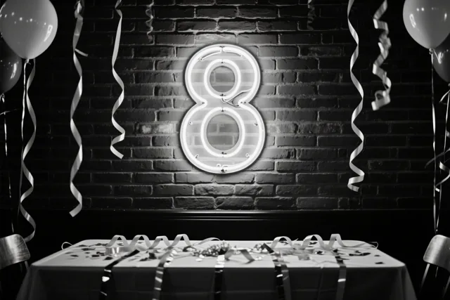Een zwart-witfoto toont een lichtgevende neonacht aan een bakstenen muur boven een feesttafel met slingers en ballonnen.