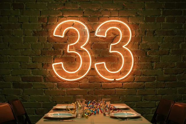 Een warm oplichtend neoncijfer 33 hangt aan een bakstenen muur boven een tafel met borden, glazen en gekleurde slingers.