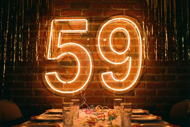 Een warm gloeiend neongetal 59 hangt aan een rode bakstenen muur boven een feesttafel met slingers en glazen.
