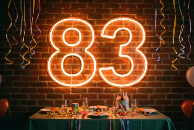 Een groot neonbord met 83 hangt tegen een bakstenen muur boven een versierde tafel met een groen tafelkleed, met kleurrijke slingers aan het plafond.