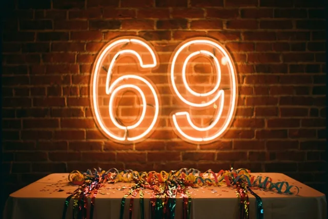 Een gloeiend neongetal 69 hangt aan een rode bakstenen muur boven een tafel met kleurrijke slingers in warm licht.