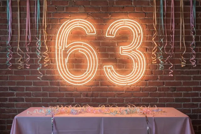 Een lichtgevend neongetal 63 hangt aan een rode bakstenen muur boven een tafel met een lichtroze tafellaken en serpentines, met kleurrijke serpentines erboven.