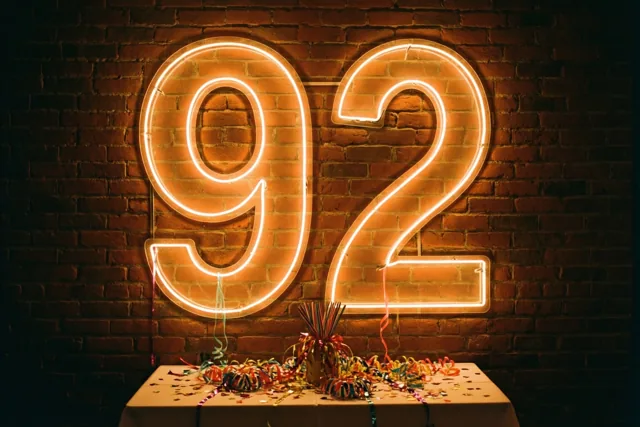 Een groot neongetal 92 gloeit op een donkere bakstenen muur boven een tafel met kleurrijke slingers en confetti.