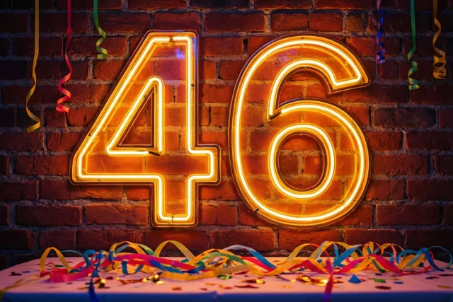 Een grote neonversie van 46 gloeit tegen een rode bakstenen muur boven een tafel met kleurrijke slingers en confetti.