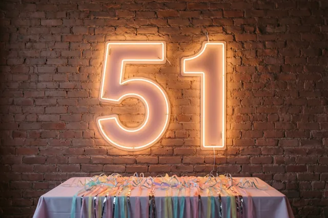 Een groot neonteken met 51 gloeit warm tegen een rode bakstenen muur boven een tafel met een lichtgekleurd kleed en kleurrijke slingers.