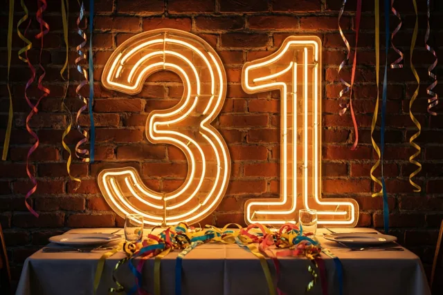 Een grote neonversiering met 31 hangt aan een rode bakstenen muur boven een tafel met een wit kleed en kleurrijke slingers.