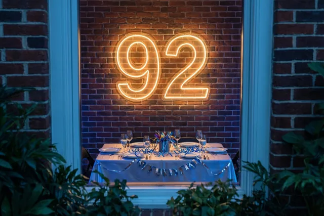 Een neongetal 92 hangt aan een bakstenen muur boven een gedekte feesttafel, gezien door een blauw omlijst raam met groen rondom.
