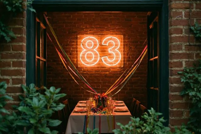Door een deuropening is een warm neonbord met 83 te zien tegen een bakstenen muur boven een feesttafel, met groen aan weerszijden en kleurrijke slingers.