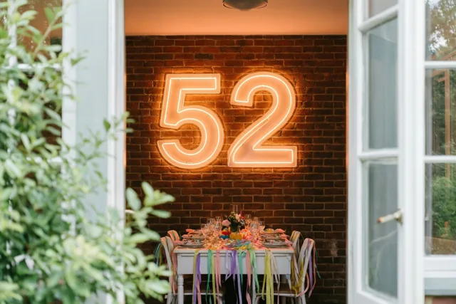 Door een open deuropening is een feesttafel met gekleurde slingers te zien onder een neonbord met 52 aan een bakstenen muur, met groen op de voorgrond.