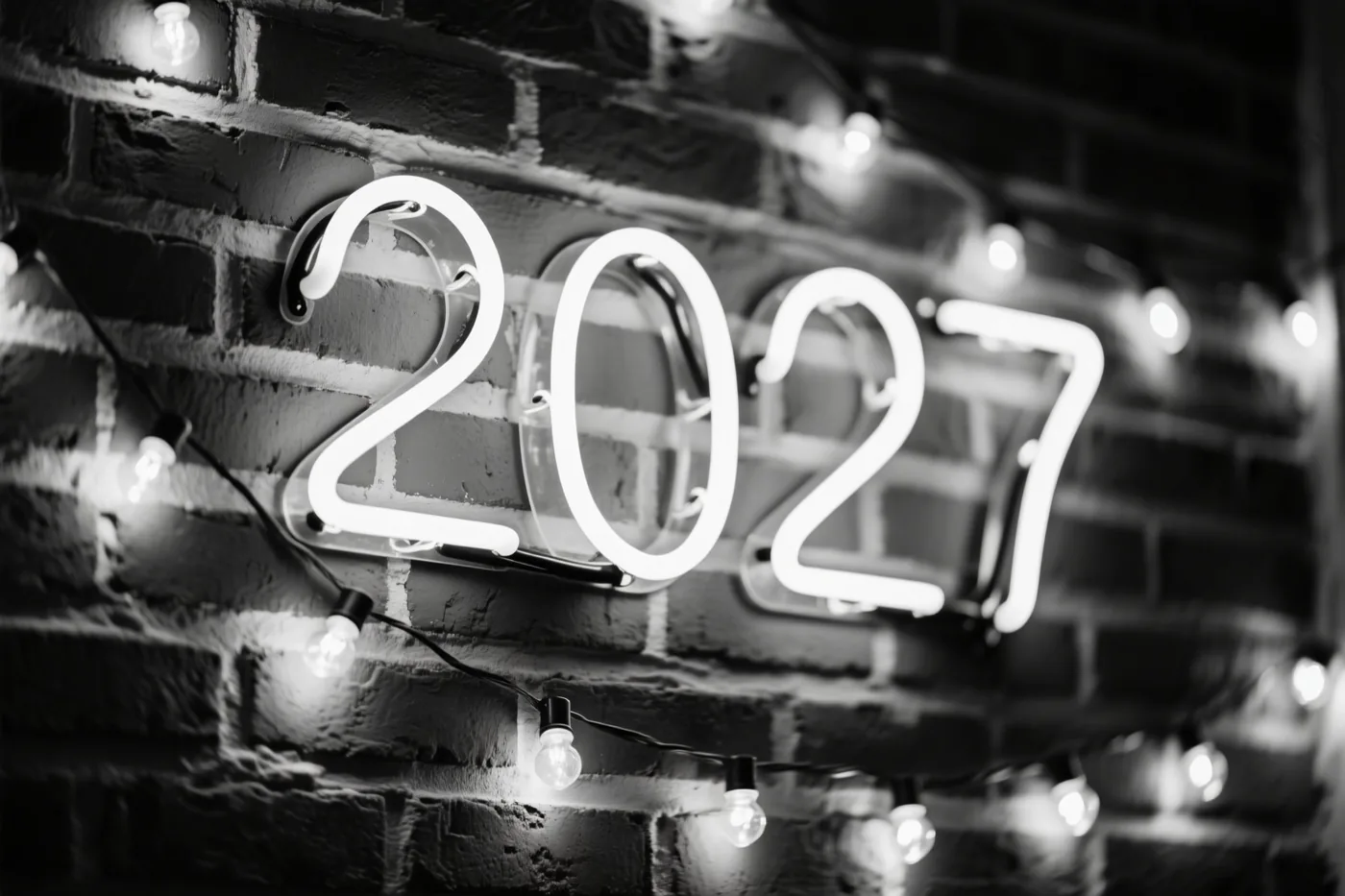 Een zwart-witfoto toont een lichtgevend neonbord met 2027 op een bakstenen muur, met lichtslingers rond het bord.