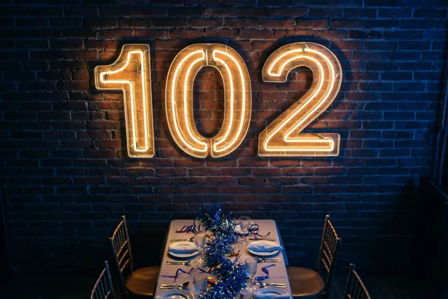 Een warm verlicht neonbord met 102 hangt aan een donkere bakstenen muur boven een feestelijk gedekte tafel met blauwe sfeerverlichting.