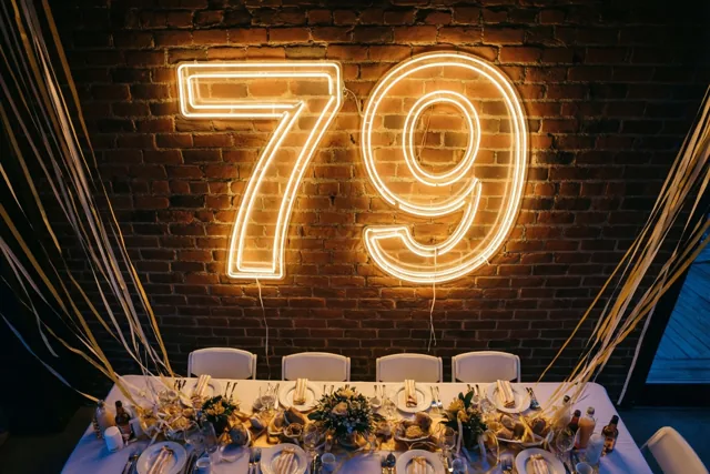 Een warm verlicht neonbord met 79 hangt aan een bakstenen muur boven een feestelijk gedekte tafel met gouden en witte slingers.